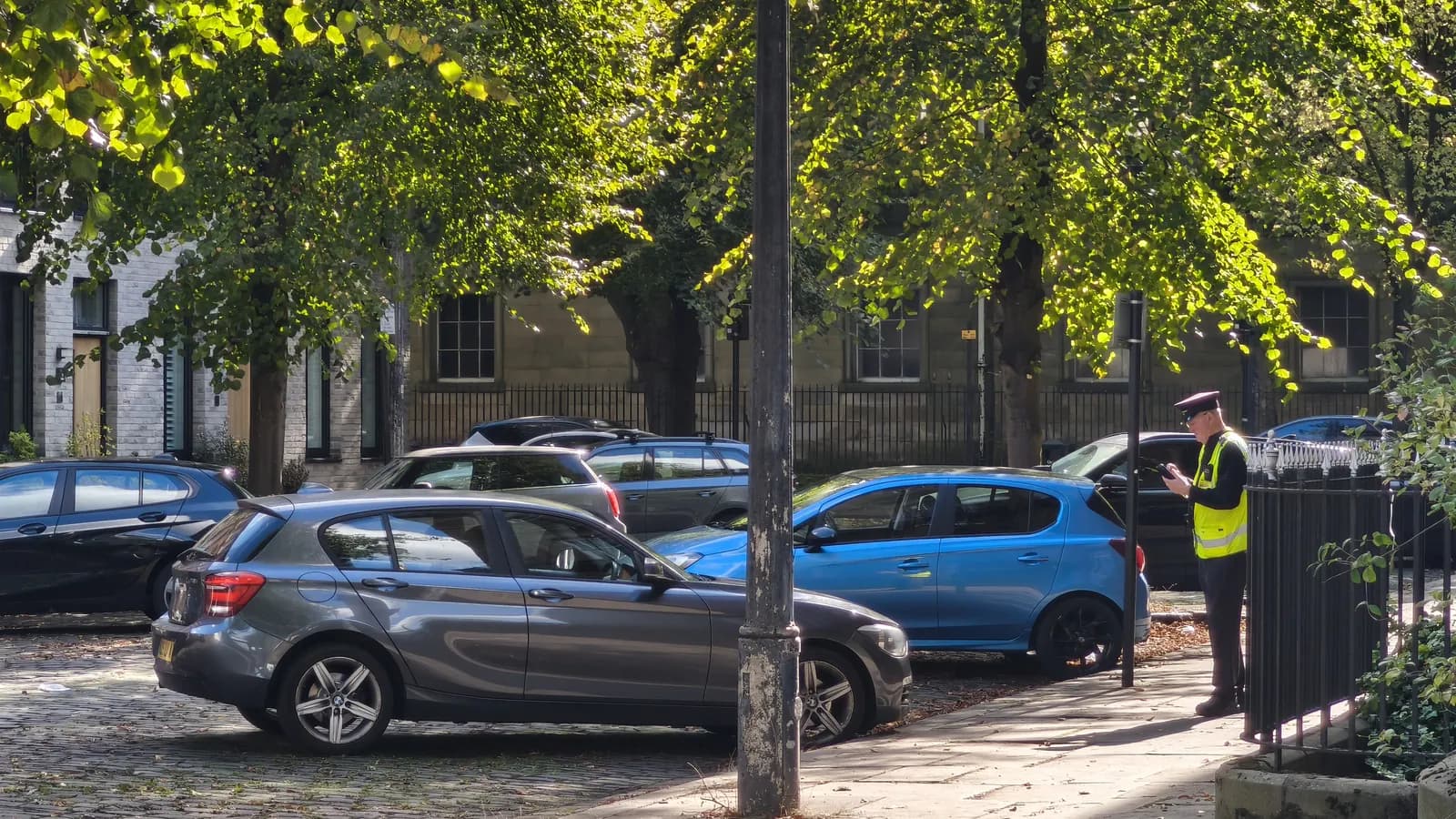 A parking enforcement officer in a high-visibility vest checking parked cars on a tree-lined street